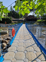 A floating bridge with a textured gray deck and blue side rails spans a shallow rocky river, with green leaves hanging from above and houses in the background.
