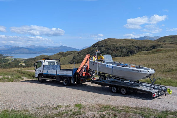 Truck with a flatbed trailer carrying a boat on a road with a scenic background.